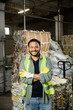 © LIGHTFIELD STUDIOS - Smiling and bearded worker in protective gloves and vest crossing arms and looking at camera while standing near blurred waste paper in waste disposal station, garbage sorting and recycling concept
