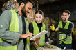 © LIGHTFIELD STUDIOS - Indian worker in protective vest using digital tablet near young colleague and sorter while working next to waste paper on hand pallet truck in waste disposal station, garbage sorting concept