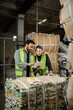 © LIGHTFIELD STUDIOS - Indian worker in safety vest using digital tablet near female colleagues while standing near waste paper on hand pallet truck in waste disposal station, garbage sorting and recycling concept