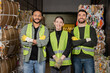© LIGHTFIELD STUDIOS - Cheerful multiethnic workers in high visibility vests and gloves crossing arms and looking at camera while standing near waste paper on disposal station, garbage sorting process