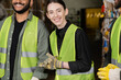 © LIGHTFIELD STUDIOS - Young worker in high visibility vest and gloves smiling at camera while standing near multiethnic colleagues in blurred waste disposal station, garbage sorting process