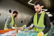 © LIGHTFIELD STUDIOS - worker in protective gloves and vest putting tin can in plastic bag while separating trash near multiethnic colleagues and conveyor in waste disposal station, garbage sorting and recycling concept