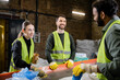 © LIGHTFIELD STUDIOS - Cheerful young woman in protective vest and gloves talking to indian colleague while separating trash near conveyor in blurred garbage sorting center, garbage sorting and recycling concept