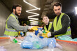 © LIGHTFIELD STUDIOS - Multiethnic sorters in protective clothes and gloves talking to colleague while working with plastic garbage on conveyor in waste disposal station, garbage sorting and recycling concept
