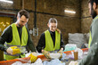 © LIGHTFIELD STUDIOS - Positive young worker in gloves and protective clothes sorting garbage on conveyor near multiethnic colleagues in waste disposal station, garbage sorting and recycling concept