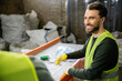 © LIGHTFIELD STUDIOS - Cheerful male sorter in protective vest and gloves looking at blurred colleague while sorting garbage near conveyor in waste disposal station, garbage sorting and recycling concept