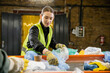 © LIGHTFIELD STUDIOS - Smiling young worker in protective vest and gloves sorting garbage near conveyor while standing in blurred waste disposal station, garbage sorting and recycling concept