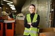 © LIGHTFIELD STUDIOS - Young and smiling worker of waste disposal station in high visibility vest looking at camera and crossing arms while standing near blurred bins at background, garbage recycling concept