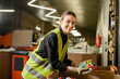 © LIGHTFIELD STUDIOS - Young and smiling female sorter in high visibility vest and gloves looking at camera while holding plastic caps near carton boxes and working in garbage sorting center, garbage sorting concept