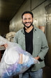 © LIGHTFIELD STUDIOS - Smiling indian volunteer looking at camera while holding plastic bag with trash in blurred waste disposal station at background, garbage sorting and recycling concept