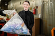 © LIGHTFIELD STUDIOS - Smiling young volunteer holding plastic bag with trash and looking at camera in blurred waste disposal station, garbage sorting and recycling concept