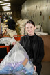 © LIGHTFIELD STUDIOS - Positive female volunteer looking at camera while holding plastic bag with trash in blurred waste disposal station at background, garbage sorting and recycling concept