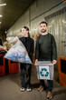 © LIGHTFIELD STUDIOS - Positive volunteers holding trash bin and bag while standing together in blurred waste disposal station at background, garbage sorting and recycling concept