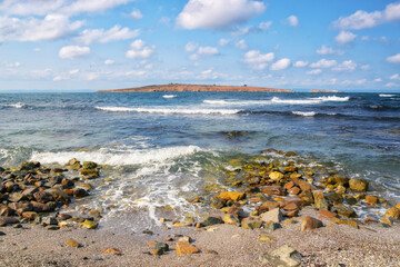 Wall Mural - wave washing stones on the shore. sunny afternoon weather with fluffy clouds on the sky above the island in the distance