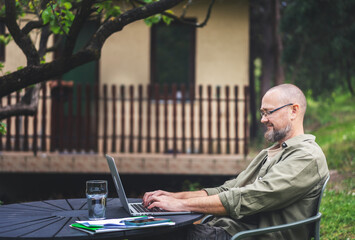 Caucasian bearded middle aged man in glasses working on project using a laptop in the backyard of a country house. Work online