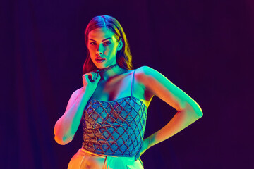 One charming woman, young fashion model with red lips wearing fashionable clothes touching necklaces posing over violet background in neon light