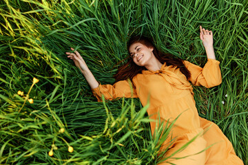 a relaxed red-haired woman enjoys summer lying in the tall green grass in a long orange dress smiling happily with her eyes closed