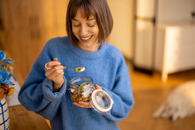 Happy Woman Enjoys Granola Breakfast In Bowl, Standing In Modern Kitchen At Home. Concept Of Wellness, Trendy Breakfasts And Domestic Lifestyle