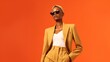 © Pixel - Beautiful young black woman posing in a trendy yellow suit, white blouse, sunglasses, and short hair. Orange background, stylish fashion photo. Empty space for text, commercial