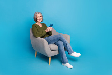 Sticker - Full length photo of satisfied pleasant woman dressed khaki sweater hold smartphone sit on armchair isolated on blue color background