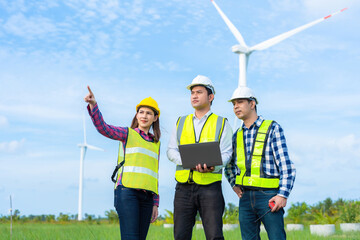 Wall Mural - Team of 3 Asian male and female engineers pointing to a destination Working people together, holding tablet, windmill turning into energy industry business, wearing vest, helmet and walkie-talkie.