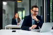 © bnenin - A businessman is working over a laptop at the office, with a female colleague in the background.