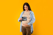 Excited Young Pregnant Woman Embracing Belly And Showing Ultrasound Picture Of Unborn Baby On Yellow Backgorund