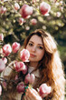 a young beautiful woman stands near the branches of a blooming pink magnolia. portrait of a girl with curly hair near a pink magnolia.