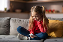 Upset Little Girl Using Mobile Phone While Relaxing On Couch At Home