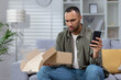 © Tetiana - A worried young Hispanic man received a wrong and damaged package, an online store order, at home. He sits at home on the sofa with the phone and contacts the post office, the delivery service