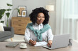 © New Africa - Happy young woman writing notes while using laptop at wooden desk indoors