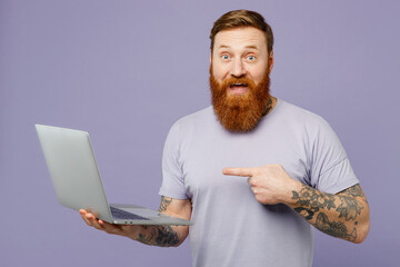 Young shocked bearded IT man wearing violet t-shirt casual clothes hold use work point finger on laptop pc computer chatting online isolated on plain pastel light purple background. Lifestyle concept.