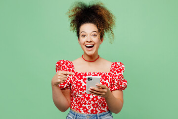 Wall Mural - Young surprised shocked happy woman she wear casual clothes red blouse hold in hand use point index finger on mobile cell phone isolated on plain pastel light green color background studio portrait