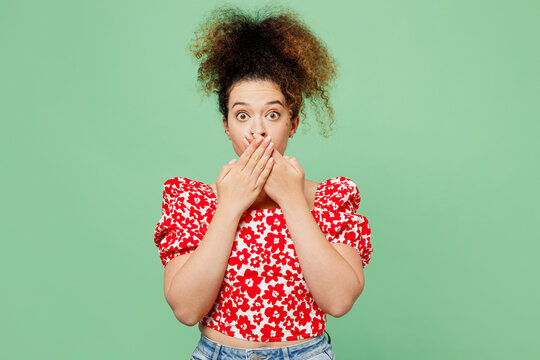Young shocked surprised sad woman she wearing casual clothes red blouse look camera covering mouth with hand isolated on plain pastel light green color background studio portrait. Lifestyle concept.