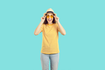 Wall Mural - Happy woman in casual clothes enjoying summer holidays. Smiling excited beautiful young girl wearing panama hat, orange sunglasses and striped tee shirt standing isolated on turquoise blue background