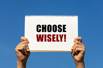 choose wisely text on notebook paper held by 2 hands with isolated blue sky background. this message