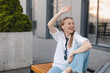 © zvkate - Happy blonde woman make hi gesture, wave her hand while meet colleagues near office building, sitting on bench and listen music in wireless earbuds. Goodbye or hello gesture of business woman.