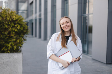 Blonde haired business lady is embracing and holding a laptop, mobile and book while posing in front of office building. Dreaming business worker walking outdoor.