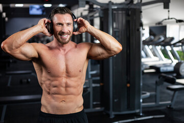  Strong sexy muscular bodybuilder guy standing  in a gym with headphones and big smile . Concept gym, crossfit, bodybuilding.