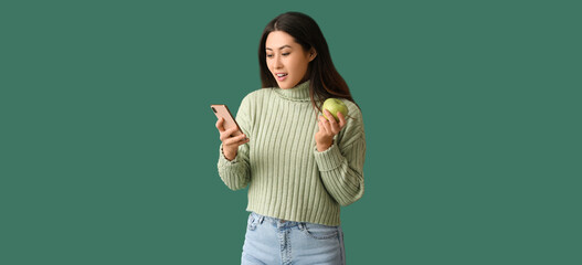 Sticker - Surprised young Asian woman with mobile phone and apple on green background