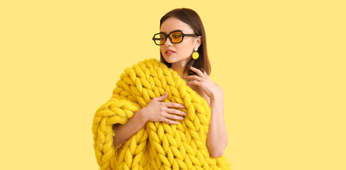 Wall Mural - Stylish young woman with knitted plaid and sunglasses on light yellow background