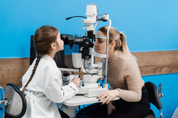 Wall Mural - Child doctor ophthalmologist with slit lamp in medical coat. Child is looking through slit lamp and examines eyes and cornea of  patient. Creative concept of ophthalmology.