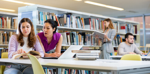 Wall Mural - Two positive female colleagues working together on joint project in library, looking for information in books and Internet..