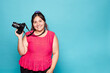 © Lux Images - Young Curvy Latina Woman Smiling, Holding a Reflex Camera, Studio Shot on Blue Background.