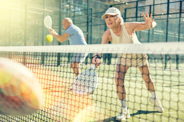 Sportive elderly man and woman engaged in Padel Tennis in open-air court of tennis club