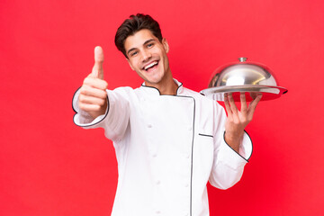 Wall Mural - Young caucasian chef man holding tray isolated on red background with thumbs up because something good has happened