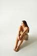 © LIGHTFIELD STUDIOS - full length of barefoot and confident woman with plus size body in strapless top with bare shoulders and underwear posing while sitting in studio on grey background, body positive, figure type