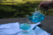 Anchan blue tea in a transparent teapot at a summer picnic in the park, next to the teapot, fruits, apricots and artichokes. and a man's hand pours tea from a teapot into a transparent bowl