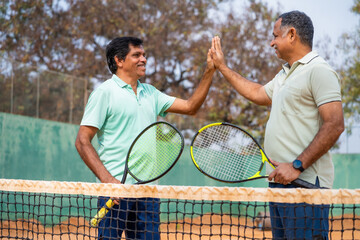 Indian two senior players celebrating by giving Hi-fi after winning tennis game at court - concept of entertainment, competition and fit or healthy retirement lifestyle