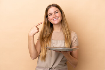 Wall Mural - Young blonde waitress woman with tray isolated on beige background giving a thumbs up gesture